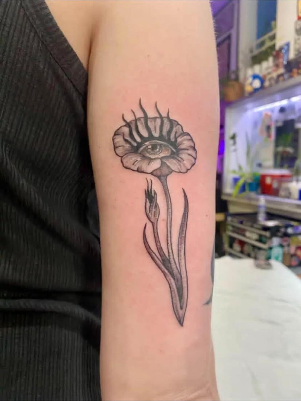 A black and gray fine line tattoo of a poppy with an eye at its center, on an upper arm